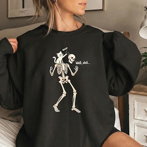 May include: A black long-sleeve sweatshirt featuring a graphic of a white cat booping the head of a skeleton. The skeleton is missing its skull, which is held in its hand. The text reads "BOOP" above the cat and "Well, shit..." next to it.