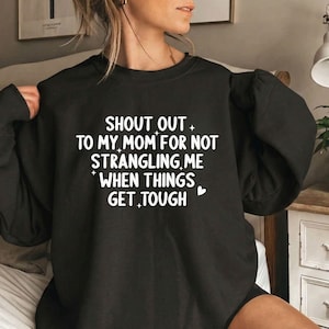 May include: A black long-sleeve sweatshirt featuring white text that reads: SHOUT OUT TO MY MOM FOR NOT STRANGLING ME WHEN THINGS GET TOUGH, accented with small white star and heart icons printed on the front center of the garment.