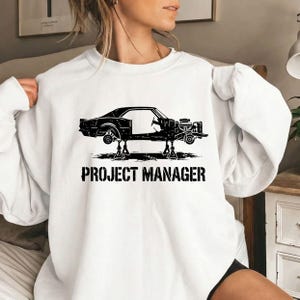 May include: A white long-sleeve sweatshirt featuring a black graphic of a car chassis resting on two jack stands. Below the car, the text PROJECT MANAGER is printed in a bold, distressed black font. The design is centered on the chest area.