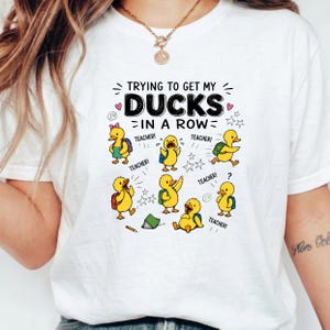 May include: A white t-shirt featuring a graphic of six yellow ducklings with backpacks and the text "TRYING TO GET MY DUCKS IN A ROW" along with the word "TEACHER!" repeated several times. The design includes small stars, hearts, and a pencil.