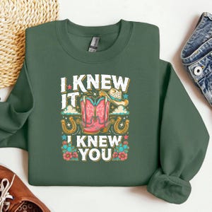 Op de afbeelding: Een donker olijfgroen sweatshirt met ronde hals en een grafische print met de tekst I KNEW IT I KNEW YOU. Het ontwerp bevat roze cowboylaarzen, een beige cowboyhoed, gouden hoefijzers, blauwe sterren, groene cactussen en rode bloemen.
