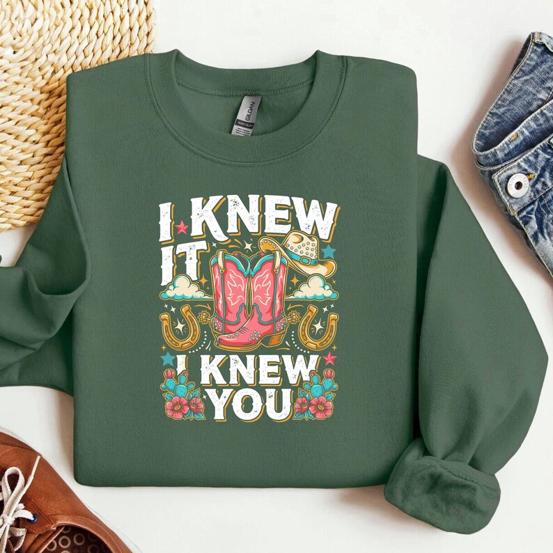 Op de afbeelding: Een donker olijfgroen sweatshirt met ronde hals en een grafische print met de tekst I KNEW IT I KNEW YOU. Het ontwerp bevat roze cowboylaarzen, een beige cowboyhoed, gouden hoefijzers, blauwe sterren, groene cactussen en rode bloemen.