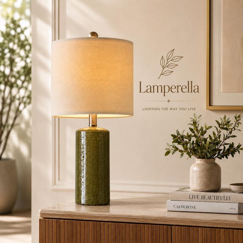 May include: A table lamp with a cylindrical olive green textured base and a beige fabric drum shade. The lamp is positioned next to a small potted plant and two stacked books with the text "LIVE BEAUTIFULLY" and "CALDERONE" on the spines.