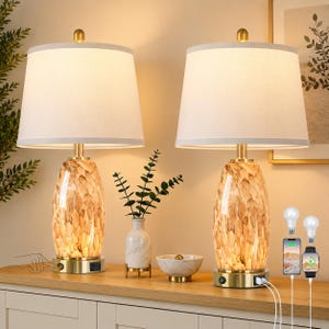 May include: A set of two table lamps featuring off-white fabric drum shades and oval glass bases with a brown and tan mottled pattern. The brass-toned metal bases include integrated USB ports and power switches for charging electronic devices.