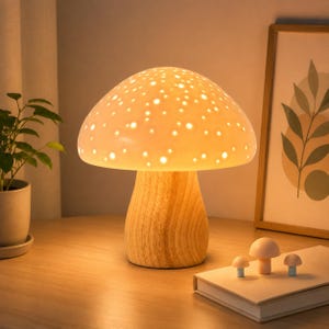 May include: A mushroom-shaped table lamp with a white, perforated ceramic cap that emits a warm glow, resting on a cylindrical wooden base with a visible grain pattern. Three small, solid mushroom figurines sit on a nearby book.