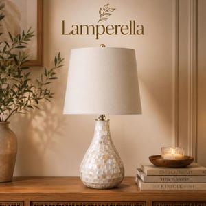 May include: A table lamp with a pear-shaped base covered in iridescent white mother-of-pearl tiles and a tapered off-white fabric shade. The brand name Lamperella is printed above the lamp. The lamp sits on a wooden surface near books.