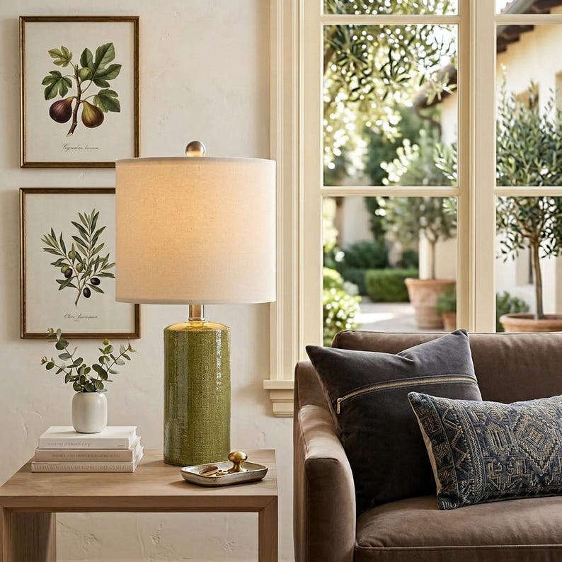 May include: A table lamp with a cylindrical olive green ceramic base and a white drum shade. The lamp sits on a wooden table next to a small white vase with green leaves, two stacked books, and a small metallic tray with a gold-toned knob.