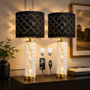 May include: Two matching table lamps with black shades featuring a gold diamond pattern. The lamps have a gold base and a clear, textured glass body. Two light bulbs, a phone, and a smartwatch are also visible.
