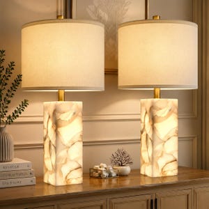 May include: A pair of table lamps featuring rectangular bases made of translucent, marbled white stone with gold-toned metal accents. Each lamp is topped with a cylindrical, off-white fabric drum shade that emits a warm, soft glow.