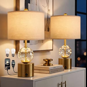 May include: Two table lamps with cylindrical beige fabric shades and brushed gold metal bases. Each lamp features a clear glass sphere containing warm LED string lights. The bases include a power switch and integrated charging ports for devices.