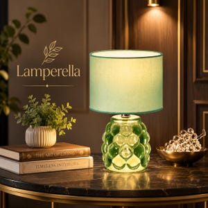 May include: A table lamp with a textured green glass base and a cylindrical light green fabric shade. The lamp is illuminated, casting a soft glow. The text Lamperella is visible in the upper left corner above two stacked books.