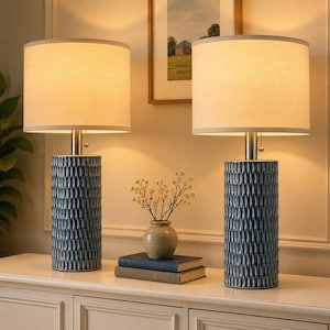 May include: Two matching table lamps with navy blue ceramic bases and beige fabric shades. The bases have a textured pattern. The lamps are on a white cabinet, with a small vase and books.