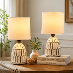 May include: Two cream-colored table lamps with beige fabric shades. The lamp bases have a geometric design with brown accents. The lamps are on a wooden table, with books and a vase nearby. The lamps are a decorative home decor item.