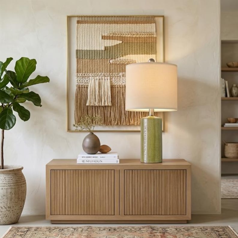 May include: A light green cylindrical ceramic table lamp with a white drum shade, positioned on a light wood cabinet with vertical slats. Behind the lamp is a framed woven wall hanging featuring cream, tan, and sage green horizontal patterns.
