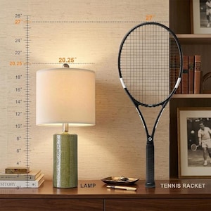 May include: A table lamp with a cylindrical green textured base and a white drum shade, standing 20.25 inches tall, positioned next to a black tennis racket that measures 27 inches in total length, as indicated by the visible measurement scale.