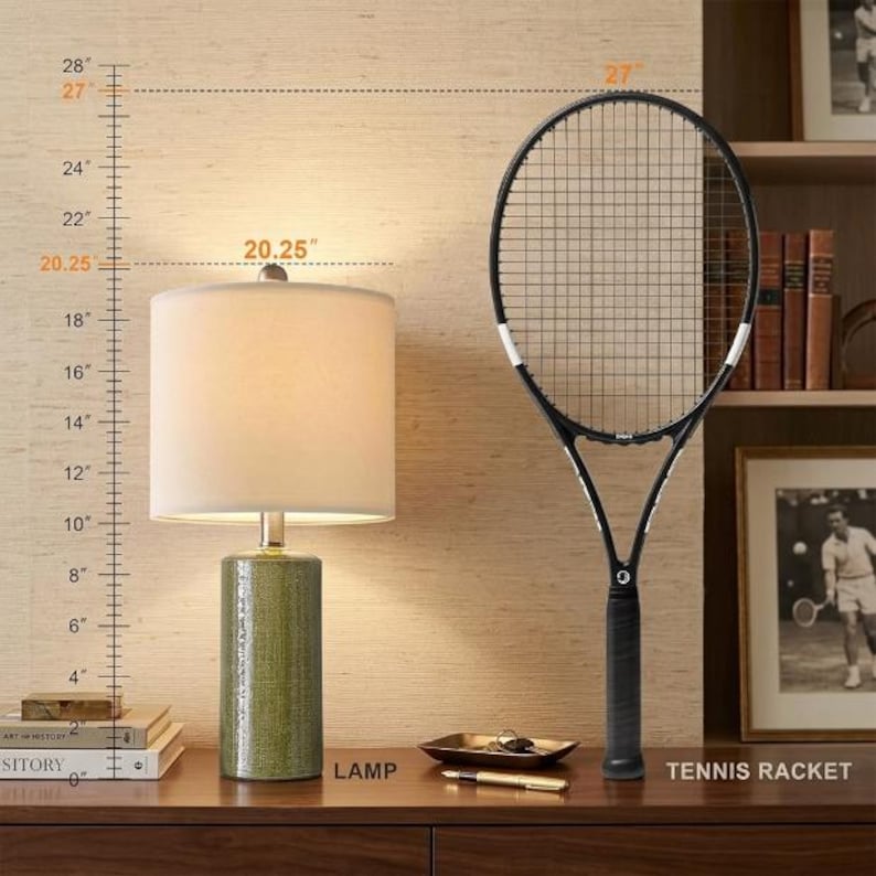 May include: A table lamp with a cylindrical green textured base and a white drum shade, standing 20.25 inches tall, positioned next to a black tennis racket that measures 27 inches in total length, as indicated by the visible measurement scale.