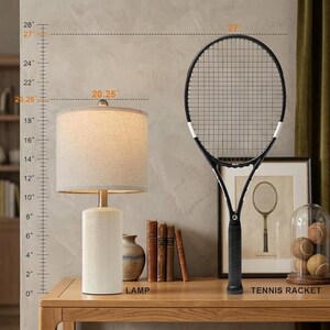 May include: A table lamp with a cylindrical white base and a beige fabric drum shade, standing 20.25 inches tall, next to a black tennis racket measuring 27 inches in height. A ruler graphic on the left indicates these specific vertical dimensions.