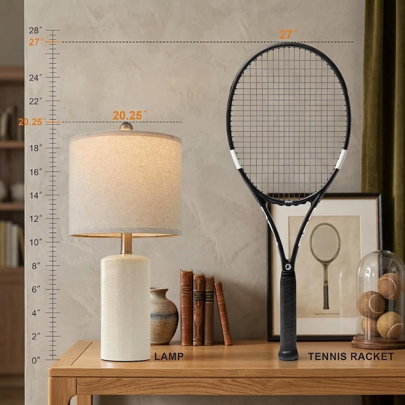 May include: A table lamp with a cylindrical white base and a beige fabric drum shade, standing 20.25 inches tall, next to a black tennis racket measuring 27 inches in height. A ruler graphic on the left indicates these specific vertical dimensions.