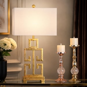 May include: A gold-colored table lamp with a rectangular white lampshade. The lamp base has a geometric design. Two candle holders with lit candles are on the right. A black vase with white flowers is on the left.