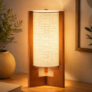 May include: A wooden table lamp with a rectangular frame and a cylindrical, off-white fabric shade. The lamp has a warm, inviting glow, and is set on a wooden surface. The lamp's design is modern and minimalist.