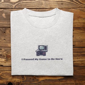May include: Light gray t-shirt with a retro video game console and controller graphic. The text "I Paused My Game to Be Here" is printed below the graphic. The shirt is folded on a wooden surface.