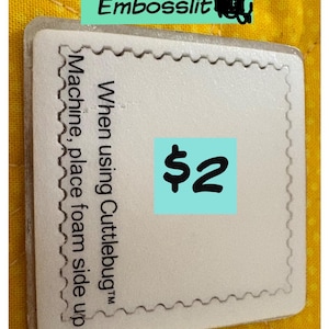 May include: White foam square with a scalloped edge. The text "When using Cuttlebug™ Machine, place foam side up" is printed on the square. A blue square with the price $2 is in the center of the foam square.