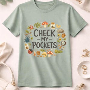 Peut inclure: T-shirt vert clair avec l'inscription "CHECK -MY- POCKETS" en gris foncé. Le texte est entouré d'une couronne d'illustrations de champignons, d'insectes et d'autres éléments naturels. Le t-shirt est en tissu doux.