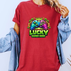 Peut inclure: T-shirt rouge avec un graphisme coloré d'une manette de jeu vidéo, de trèfles et d'un fer à cheval. Le texte "LUCKY GAMER MODE" est affiché en dessous de la manette en vert et blanc. Le t-shirt est de style décontracté.