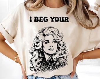 May include: A cream-colored t-shirt featuring a black graphic print of a woman with voluminous hair and hoop earrings. Above the illustration, the text reads I BEG YOUR in a bold, rounded font. The design includes a rose detail on the shoulder.