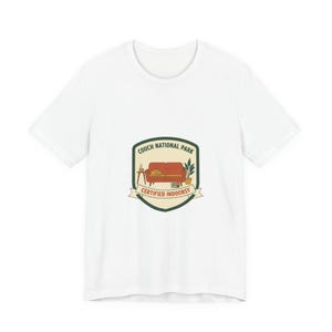 Op de afbeelding: Wit T-shirt met een grafisch ontwerp. Het ontwerp bevat de tekst "Couch National Park" en "Certified Indoorsy" met een illustratie van een bank, tafel en planten. Het ontwerp heeft de vorm van een schild.