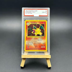 Puede incluir: Una carta de Pokémon de 1999, Charizard Holo 1st Edition, calificada. La carta presenta una ilustración de Charizard amarillo con detalles en rojo y naranja. La carta está en un soporte protector y sobre un caballete de madera.