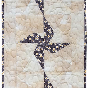 May include: A rectangular quilted table runner with a repeating geometric pattern. The design features alternating sections of beige and dark gray fabric with white floral accents. The edges are finished with a dark gray border.