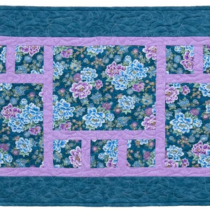 May include: A rectangular quilt with a teal border and a floral center. The center features a dark blue background with blue and purple flowers. A lavender frame surrounds the floral pattern. The quilt is likely handmade.