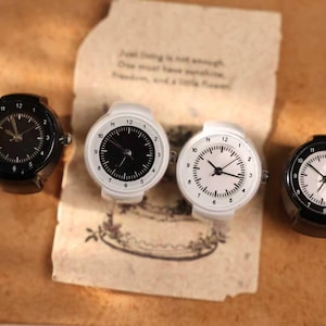 Può includere: Quattro anelli orologio con quadranti rotondi. Due sono neri con segni bianchi e due sono bianchi con segni neri. Gli orologi sono su una superficie marrone con un pezzo di carta con testo.