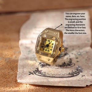 May include: A gold-colored ring shaped like a wristwatch, featuring a square face with black numerals on a yellow background. The ring has a quartz movement and the text "QUARTZ" is visible. The image includes text about engraving.