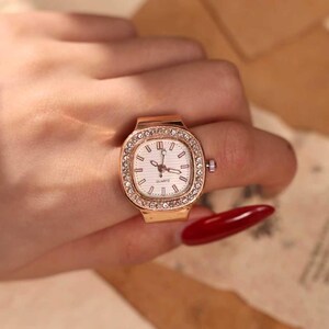 May include: A gold-toned ring watch with a square face and a white dial. The watch is surrounded by a row of small, clear crystals. The watch is worn on a finger.