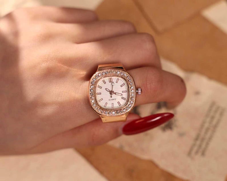 May include: A gold-toned ring watch with a square face and a white dial. The watch is surrounded by a row of small, clear crystals. The watch is worn on a finger.