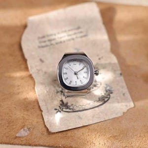 Puede incluir: Un reloj de anillo plateado con esfera cuadrada y blanca. El reloj tiene manecillas e indicadores de hora negros. El reloj est&aacute; sobre un trozo de papel envejecido con texto descolorido e ilustraciones florales.