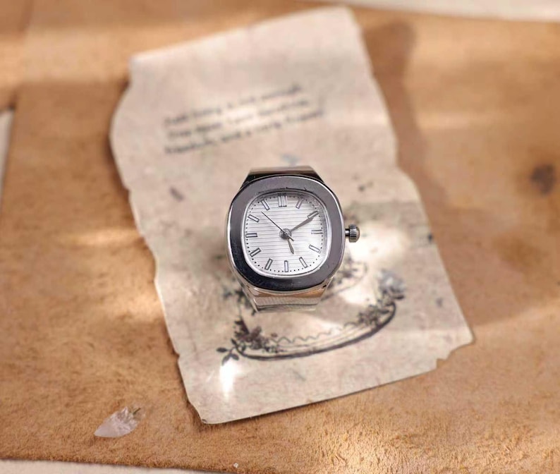 Puede incluir: Un reloj de anillo plateado con esfera cuadrada y blanca. El reloj tiene manecillas e indicadores de hora negros. El reloj est&aacute; sobre un trozo de papel envejecido con texto descolorido e ilustraciones florales.