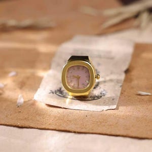 May include: A gold-toned ring watch with a square face and a pink dial. The watch has a black band and is set on a piece of aged paper. The background is a warm, neutral tone.