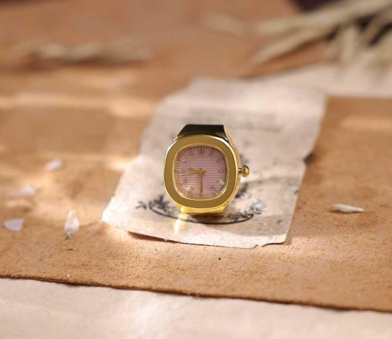 May include: A gold-toned ring watch with a square face and a pink dial. The watch has a black band and is set on a piece of aged paper. The background is a warm, neutral tone.