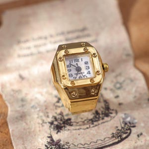 May include: A gold-toned ring watch with a square face. The watch face is white with black numerals and hands. The ring has a textured design and a small crown. The watch is set on a vintage paper.
