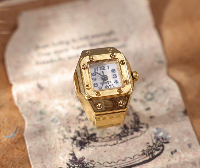 May include: A gold-toned ring watch with a square face. The watch face is white with black numerals and hands. The ring has a textured design and a small crown. The watch is set on a vintage paper.