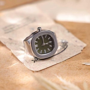 Puede incluir: Un anillo plateado con una esfera de reloj. La esfera es cuadrada con fondo verde oscuro y agujas e indicadores plateados. El anillo est&aacute; sobre un trozo de papel envejecido.