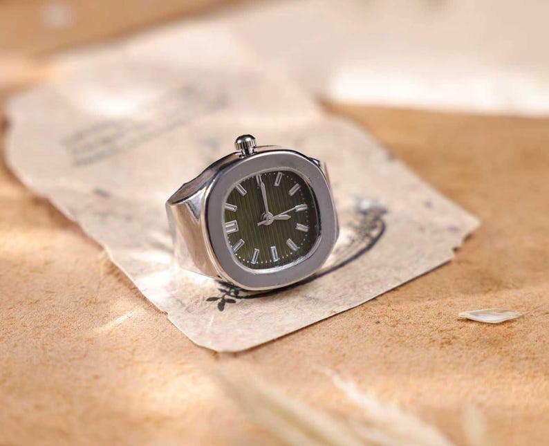 Puede incluir: Un anillo plateado con una esfera de reloj. La esfera es cuadrada con fondo verde oscuro y agujas e indicadores plateados. El anillo est&aacute; sobre un trozo de papel envejecido.