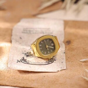May include: A gold-toned watch ring with a green watch face. The ring has a rectangular shape with rounded corners. The watch face features gold-colored hands and hour markers. The ring is resting on a piece of aged paper.