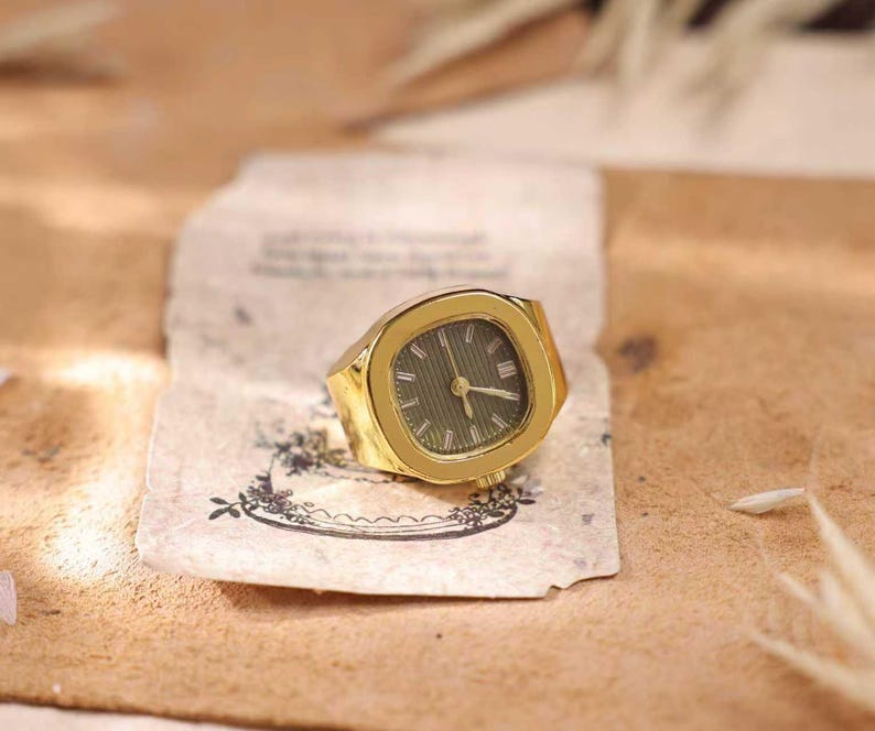 May include: A gold-toned watch ring with a green watch face. The ring has a rectangular shape with rounded corners. The watch face features gold-colored hands and hour markers. The ring is resting on a piece of aged paper.