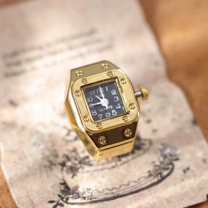 May include: A gold-toned ring watch with a square black face. The watch has a gold bezel with a small crown and a black watch face with white numerals and hands. The ring is on a vintage paper background.