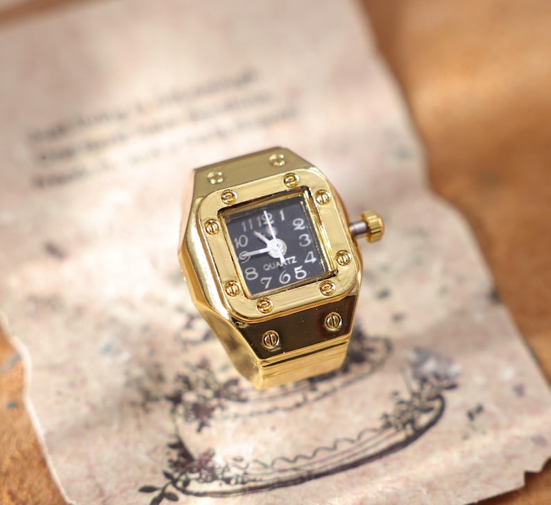 May include: A gold-toned ring watch with a square black face. The watch has a gold bezel with a small crown and a black watch face with white numerals and hands. The ring is on a vintage paper background.
