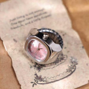 Puede incluir: Un reloj de anillo plateado con esfera rosa. El reloj tiene una esfera rectangular con una peque&ntilde;a corona. La correa es el&aacute;stica. El reloj est&aacute; sobre un papel con texto.